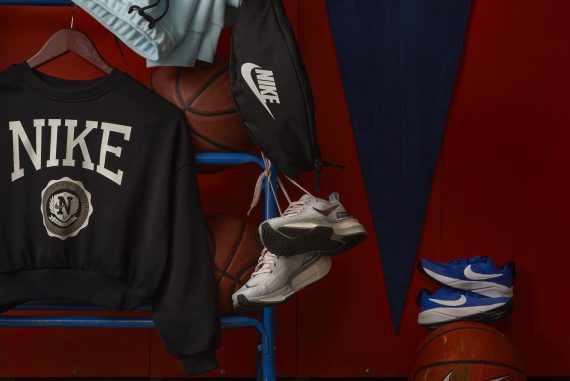 Nike gym class essentials hanging on a basketball rack with a pennant flag in the background, including a Nike crew sweatshirt, waist pack, sweatpants, three pairs of shoes and Nike basetballs